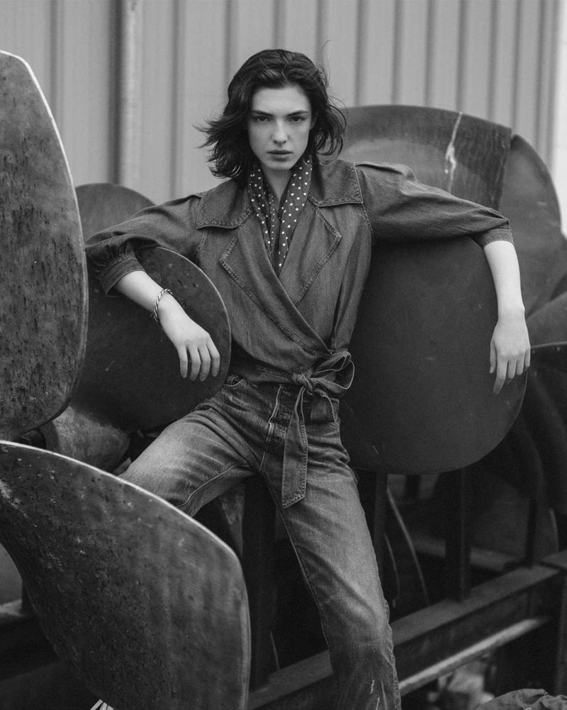 Black and white fashion portrait of a woman in denim jacket and jeans, posing with large metal objects in an outdoor setting.