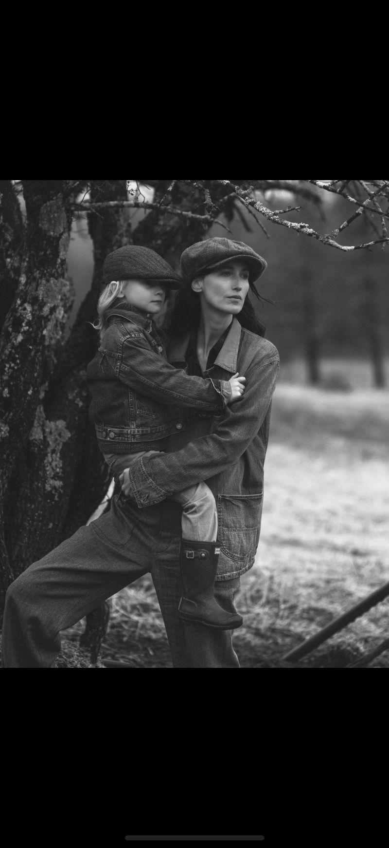 A woman and a child wearing hats and jackets are standing next to a tree in a black and white outdoor shot, looking off into the distance.