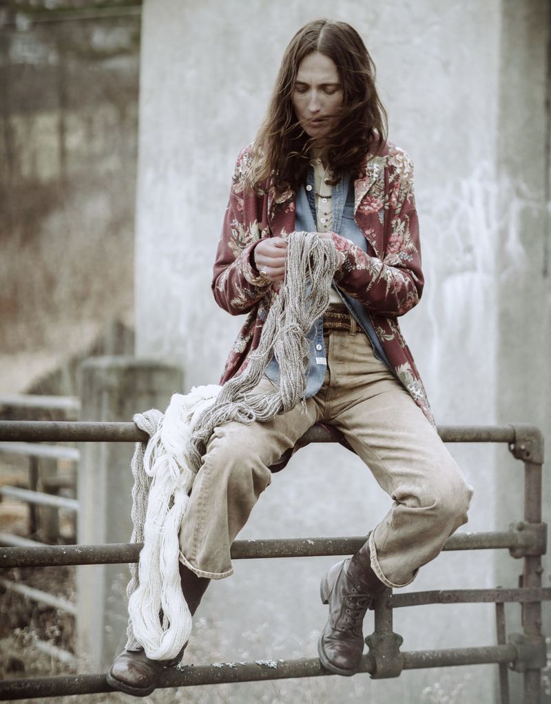 Model sitting on a metal railing outdoors, holding raw yarn while wearing a patterned floral jacket and denim shirt.