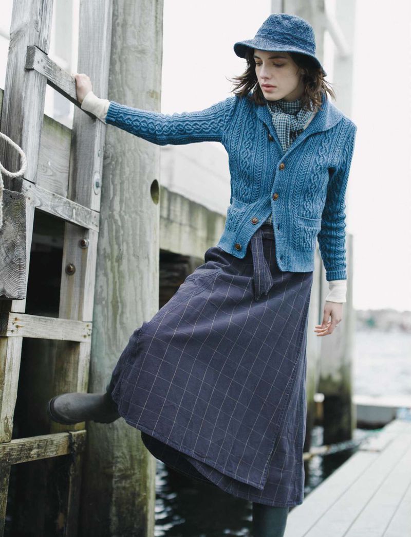 Woman in blue cardigan, plaid skirt, and hat standing on a wooden dock near water, posing in an outdoor setting during daylight.