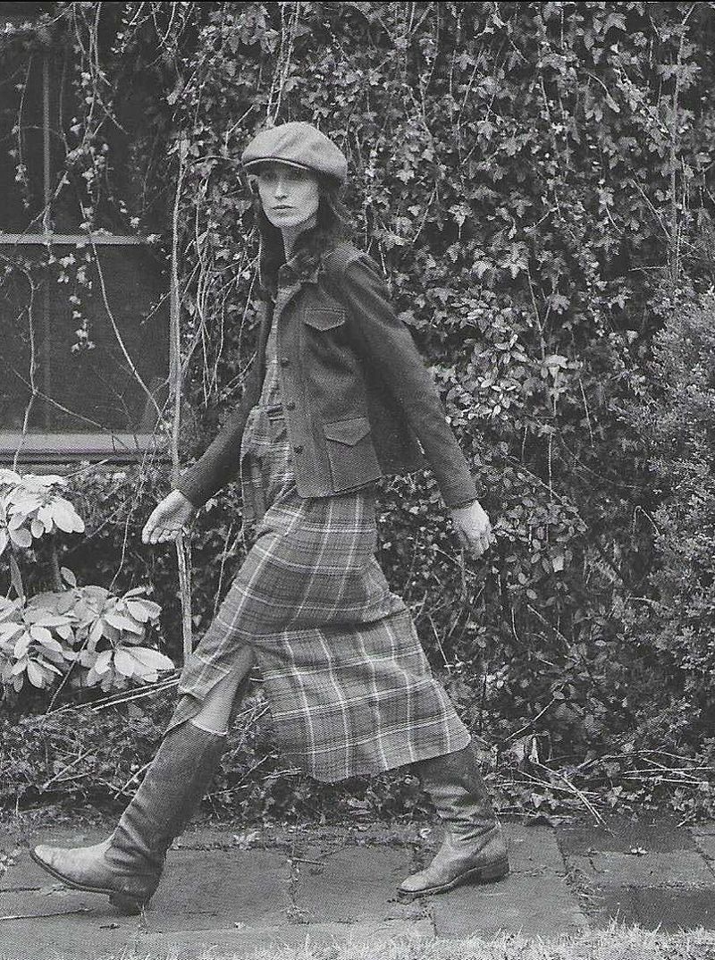 A full-length shot of a woman walking outdoors, wearing a cap, jacket, and plaid skirt with boots, with green vines in the background.