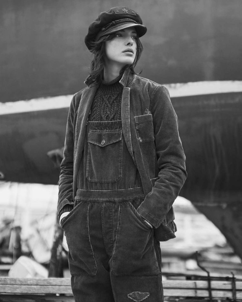 Monochrome portrait of a person wearing overalls and a cap, posing outdoors in front of a blurred backdrop with hands in pockets.