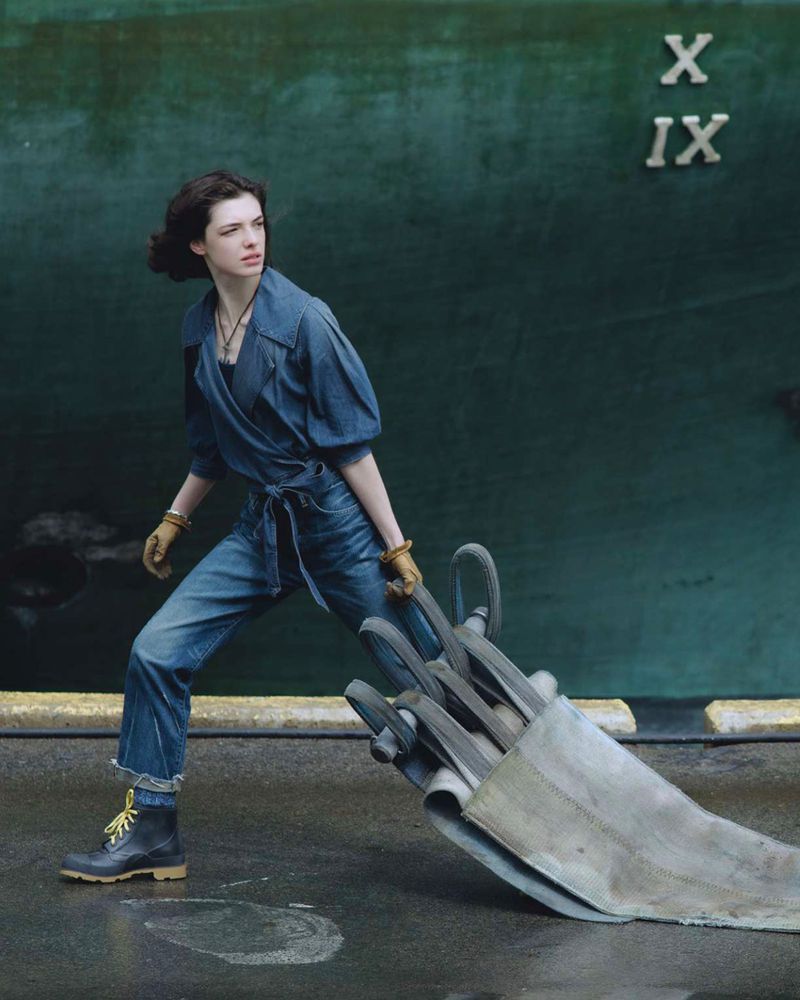 Woman in blue denim jumpsuit pulls a silver metal object on concrete against a green wall with Roman numerals. 