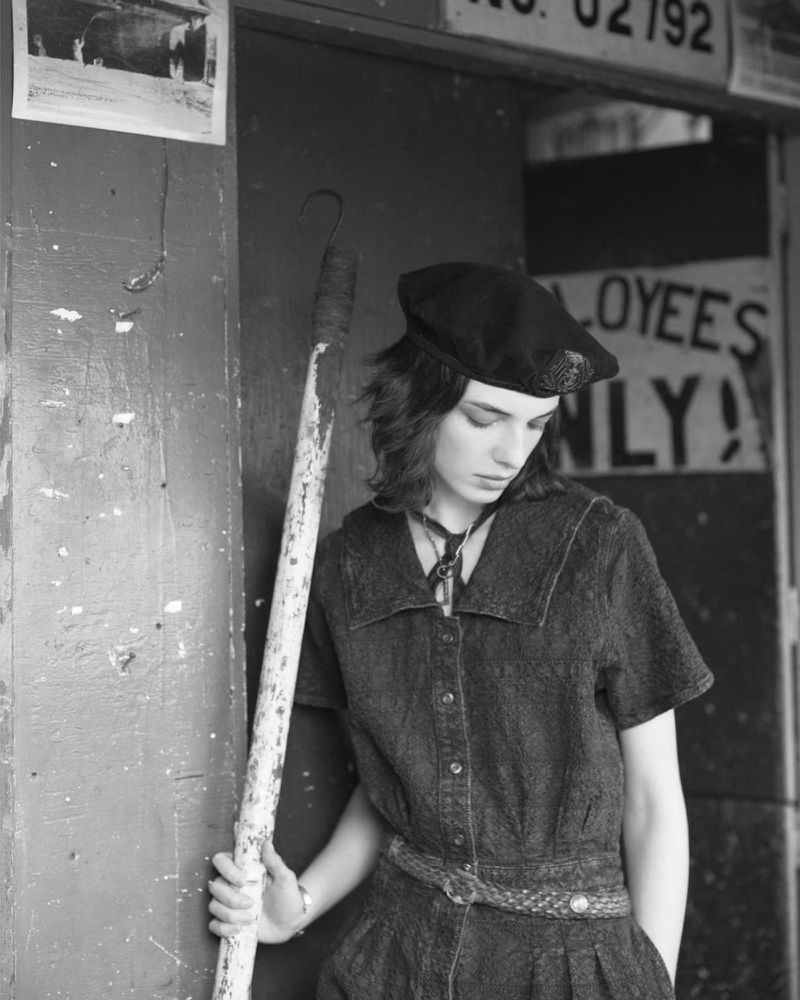 Young woman in a vintage-style denim dress and beret stands by a door, holding a pole with a hook in an urban setting in black and white.