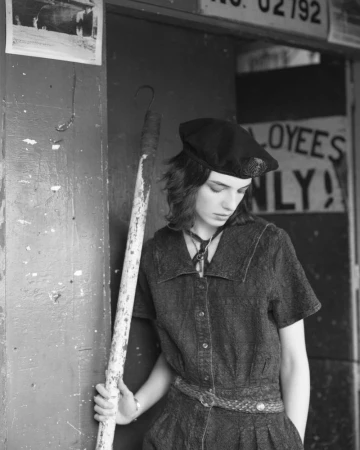 Young woman in a vintage-style denim dress and beret stands by a door, holding a pole with a hook in an urban setting in black and white.