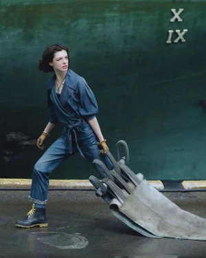 Model in denim jumpsuit and work boots dragging heavy industrial straps outdoors.