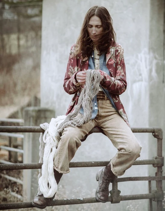 Model sitting on a metal railing outdoors, holding raw yarn while wearing a patterned floral jacket and denim shirt.