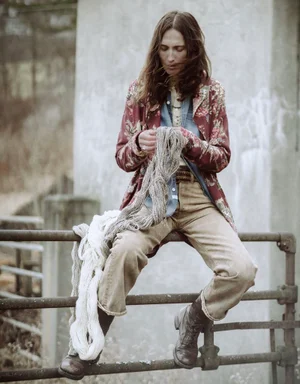 Model sitting on a metal railing outdoors, holding raw yarn while wearing a patterned floral jacket and denim shirt.