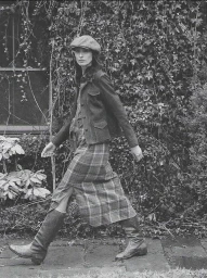 Model in flat cap and plaid midi skirt walking outdoors past dense foliage for Ralph Lauren RRL