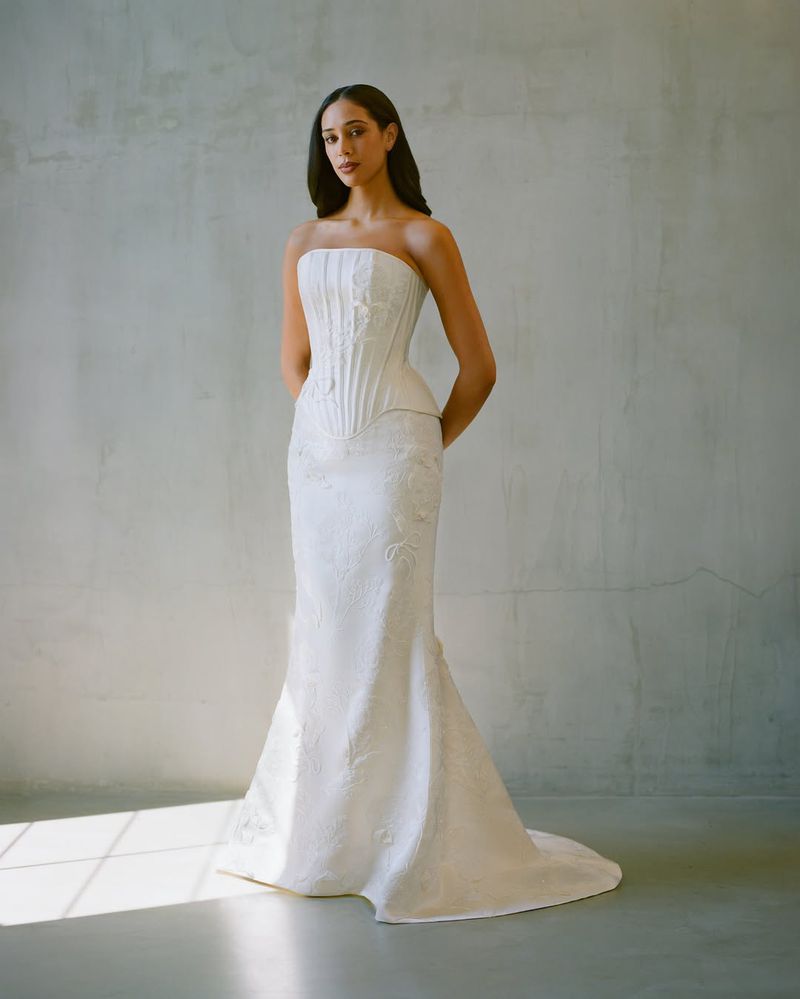 Woman in white strapless gown stands in front of neutral background, hands clasped. Formal wear, fashion portrait, full-length shot.