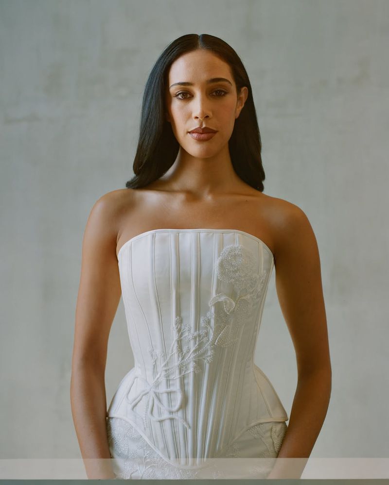 Portrait of a woman with long dark hair wearing a strapless white corset top against a neutral background in a studio setting.