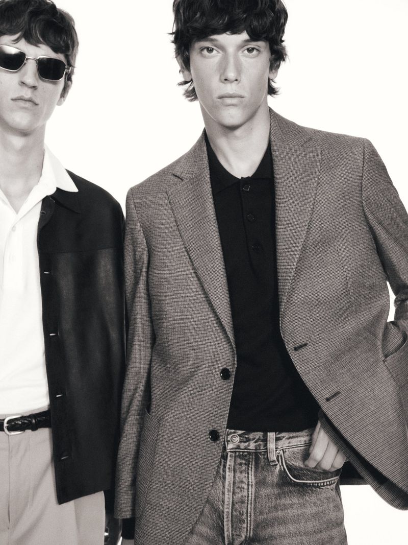 Two adult men in a monochrome studio shot. One in a suit jacket and jeans, the other in a black jacket, sunglasses, and light pants.
