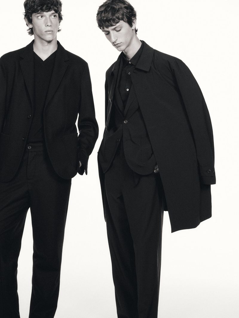 Two men in dark suits, one with a coat draped over his shoulder, stand against a white background in a studio setting.