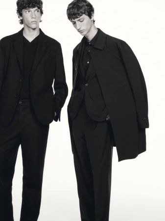 Two men in dark suits, one with a coat draped over his shoulder, stand against a white background in a studio setting.