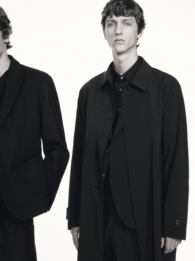 Two adult men pose indoors in black overcoats and button-down shirts against a bright white backdrop in a fashion editorial style.