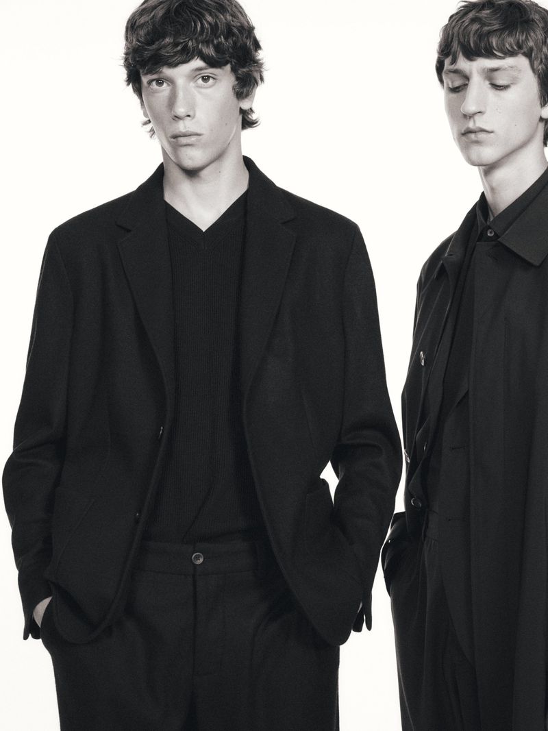 Two young male models in black blazers and pants pose against a bright white backdrop. Fashion editorial shot with a focus on menswear.
