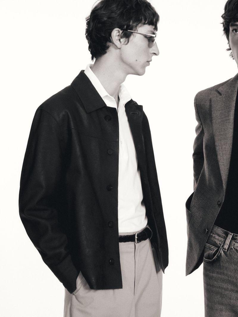 Two men wearing tailored jackets pose against a bright background in a studio shot; one wears sunglasses, a collared shirt and dress pants.