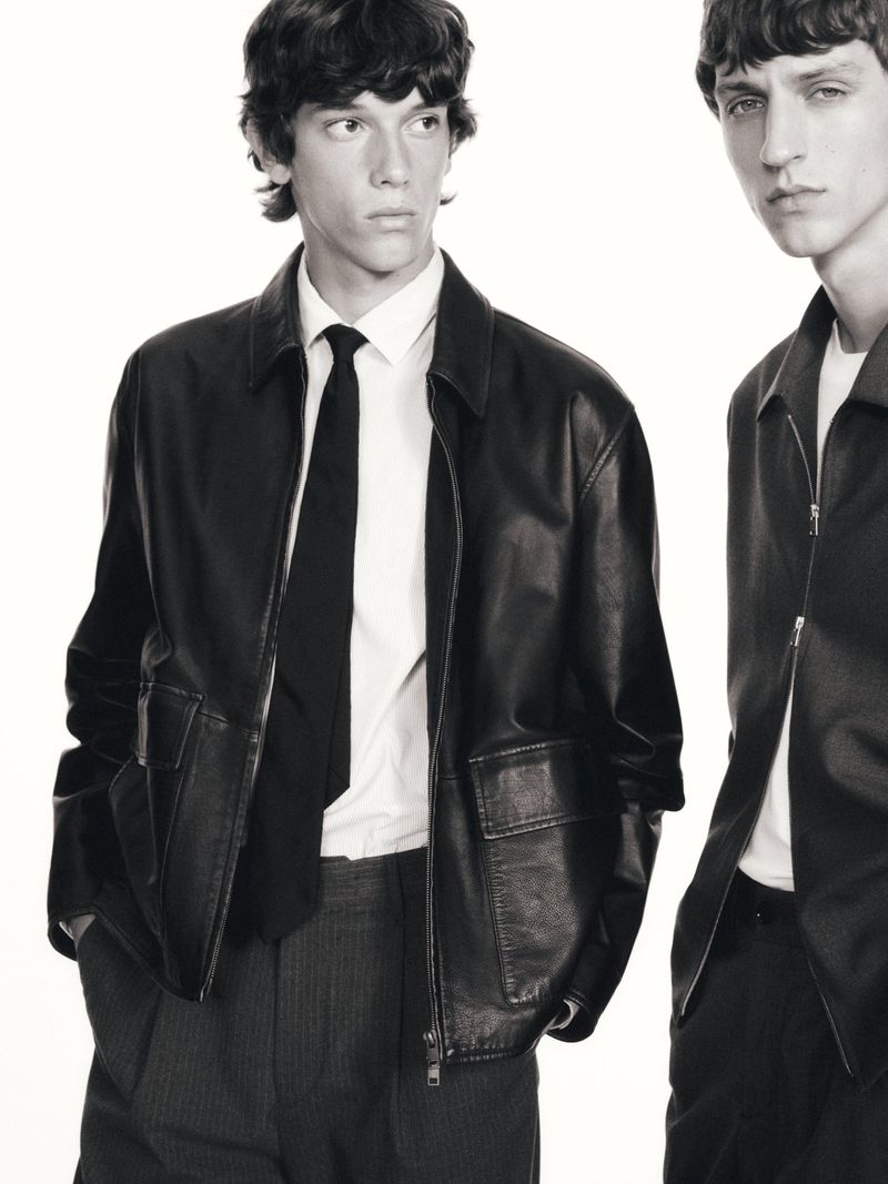 Two young adult men pose indoors wearing black leather jackets, white shirts, and dark ties against a bright white background.