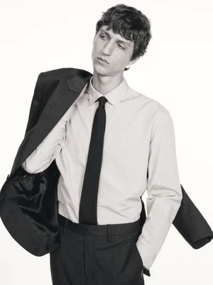 A male model wearing a button-down shirt, tie, and trousers with a jacket slung over his shoulder against a plain white background.