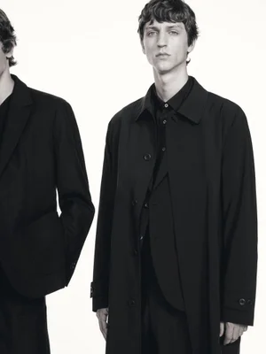 Two male models in dark black coats and shirts standing against a solid white background in a studio setting
