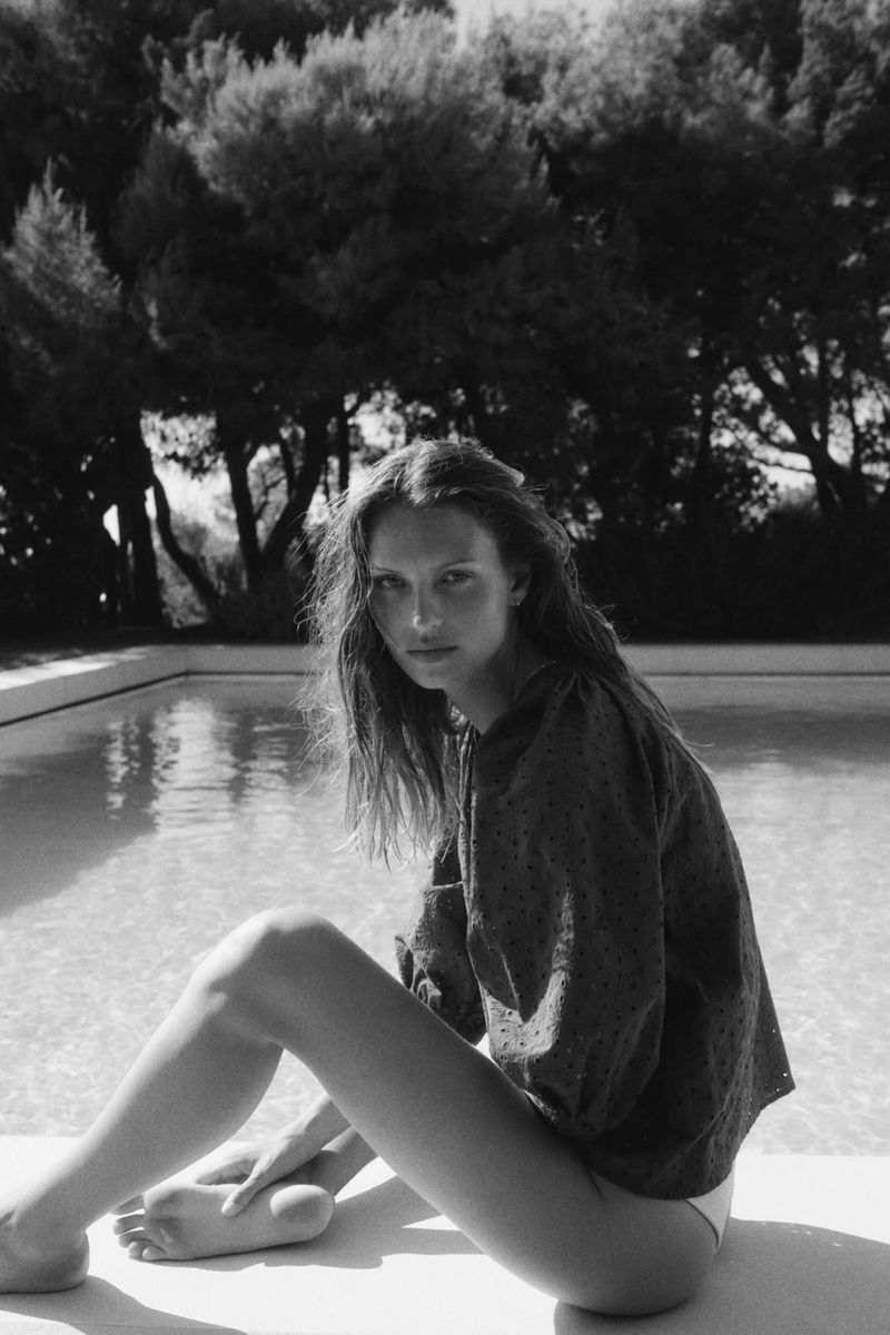 Woman in collared top sitting near the edge of a swimming pool with trees in the background in a black and white outdoor shot.
