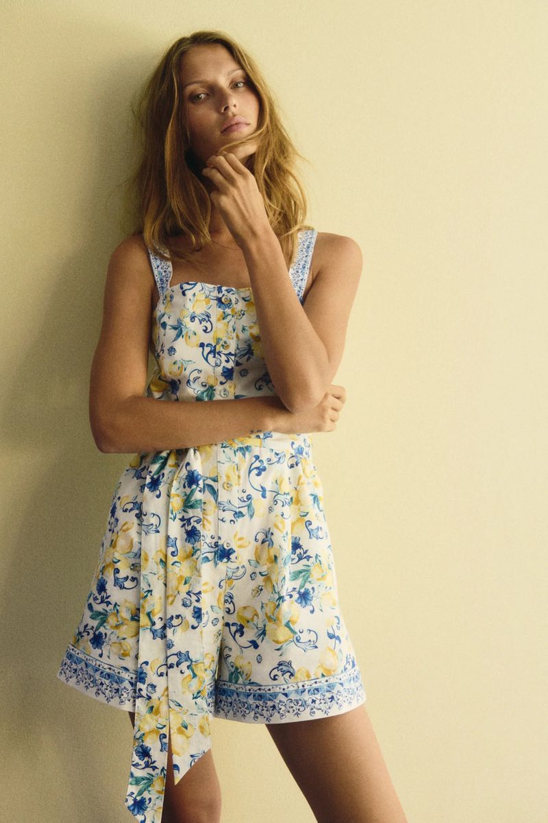 A fair-skinned adult female poses in a sleeveless romper with a white, yellow, and blue floral pattern in a studio setting.