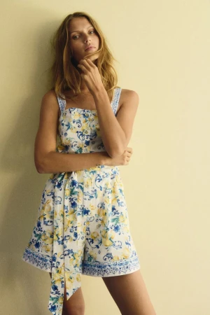 A fair-skinned adult female poses in a sleeveless romper with a white, yellow, and blue floral pattern in a studio setting.