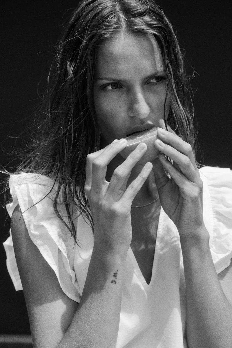 Monochrome close-up of an adult woman eating a slice of orange, wearing a white V-neck top with ruffled sleeves against a dark background.