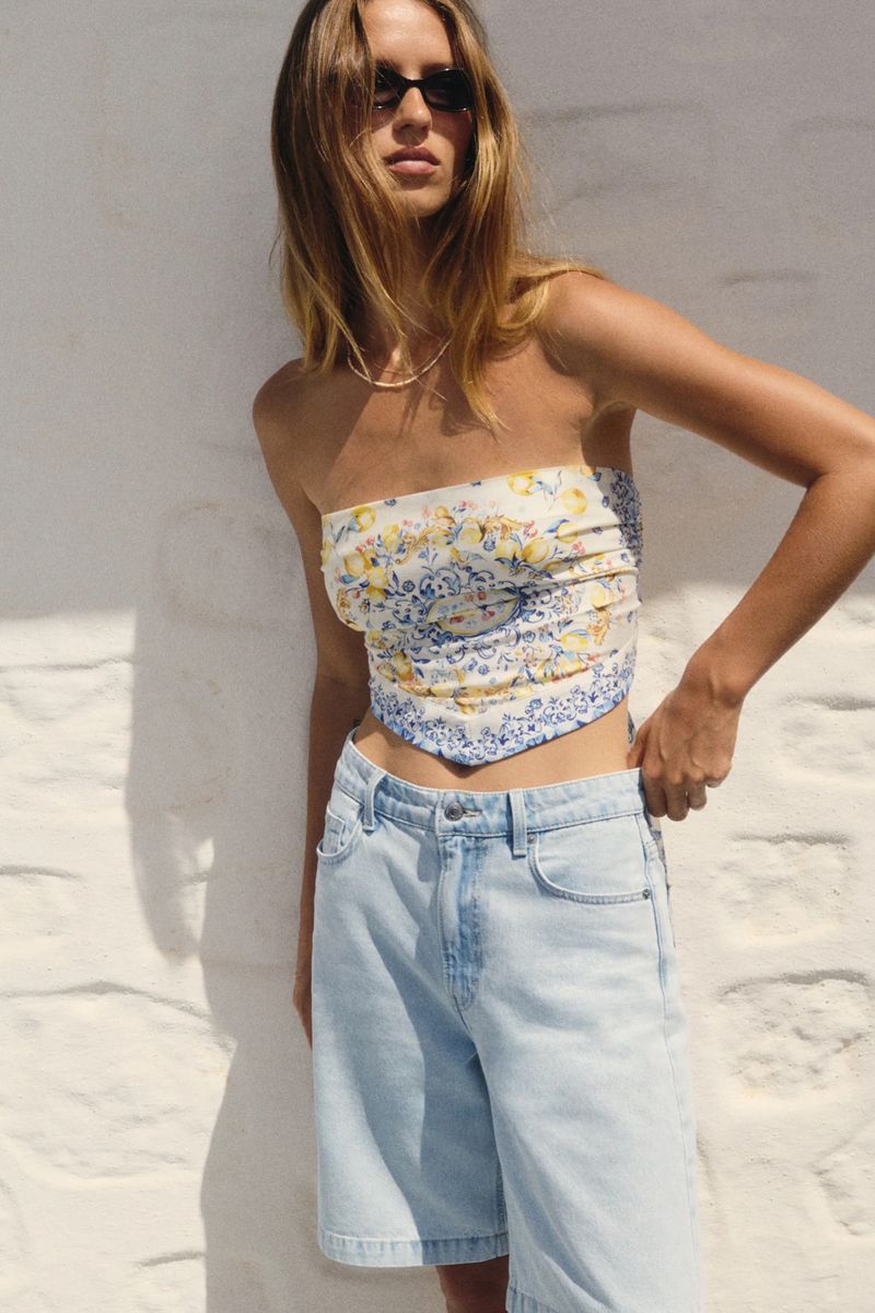 Woman in a strapless floral top and light-wash denim shorts stands against a sunlit wall, showcasing a summer fashion editorial look.
