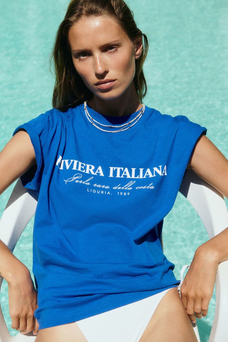 Model wearing a blue t-shirt with white text seated in a chair by an outdoor pool for the H&M Coastal Summer 2025 collection.