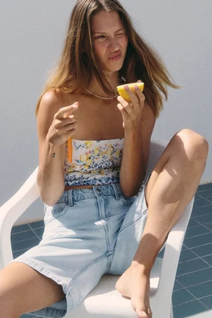 Young adult woman sits in a white chair, wearing denim shorts and a floral top, holding a halved lemon in bright, sunny outdoor setting.