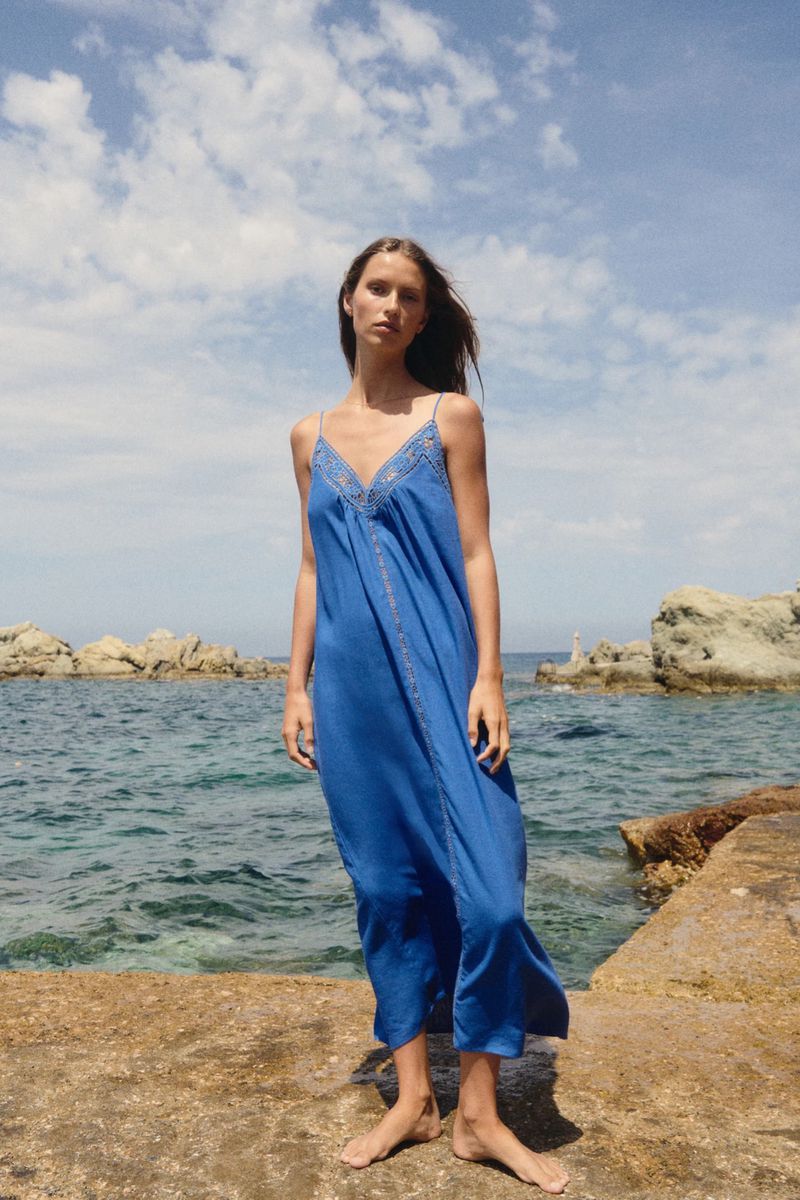 A woman stands on a stone outcrop overlooking the ocean, wearing a loose-fitting blue dress with a V-neck and thin straps.