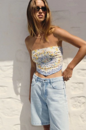 Model in dark sunglasses wearing a patterned strapless top and light denim shorts against a white wall.