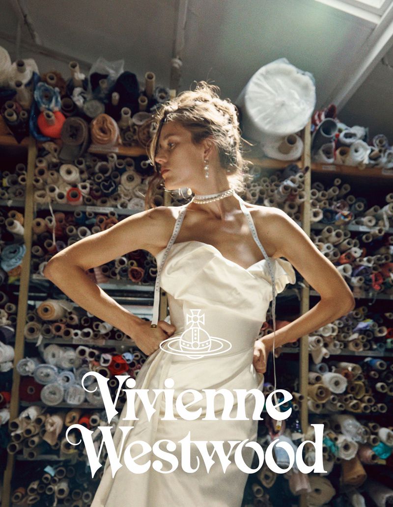 A woman with arms raised in a cream-colored dress and pearls poses in front of bolts of fabric in a fashion or design studio.