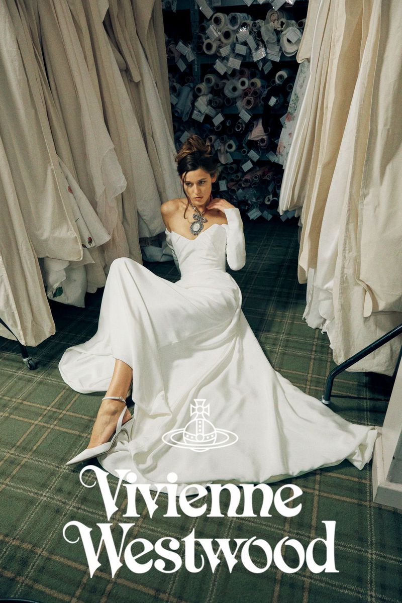 A woman in a Vivienne Westwood gown poses in what appears to be a designer's workshop surrounded by draped dresses and rolls of fabric.