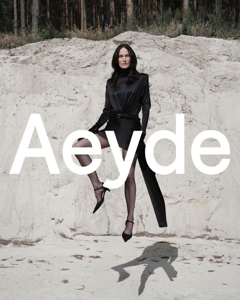 A woman in a black dress and tights poses in front of a white rock formation and a pine forest for a fashion brand advertisement.
