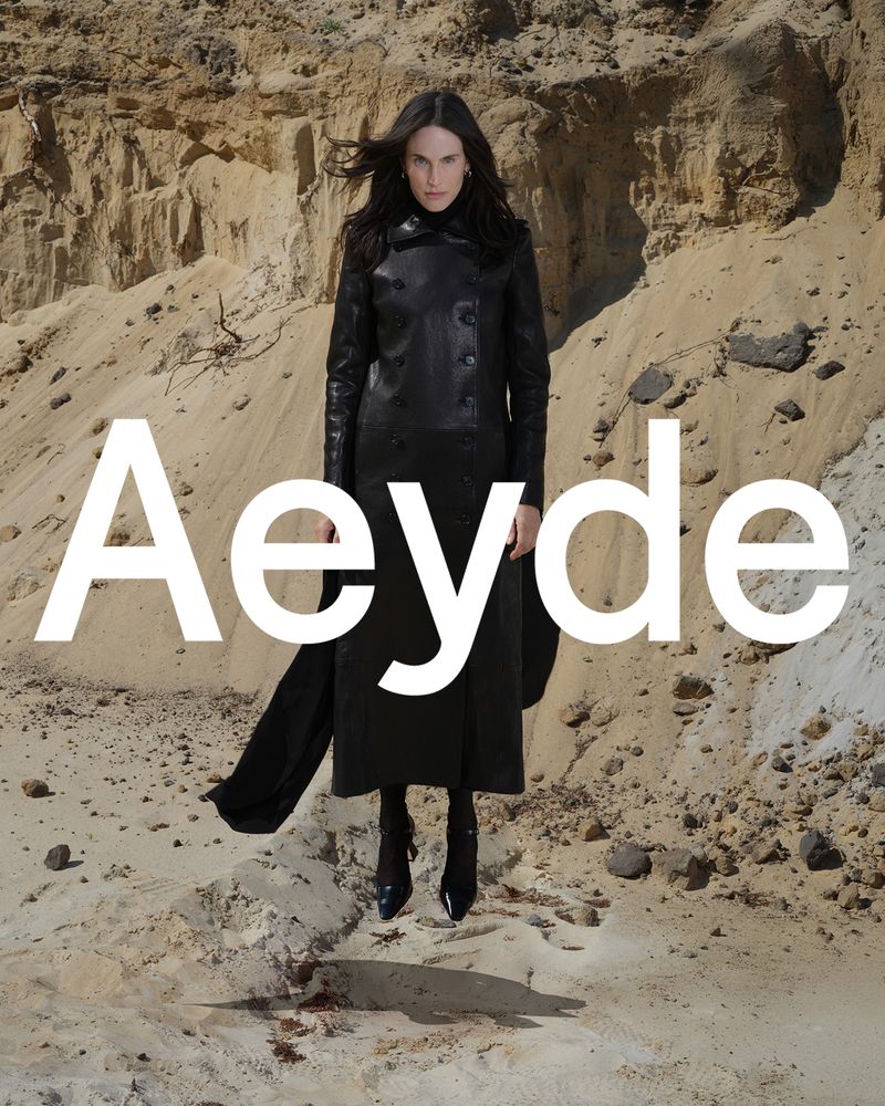 An adult female in a black coat and boots stands in a sandy, outdoor landscape under natural light. The Aeyde brand logo is visible.