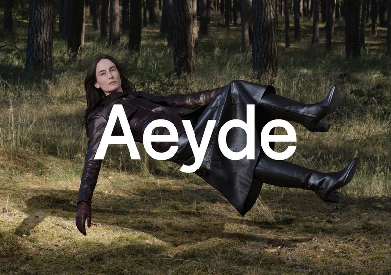 Fashion shot of woman in black leather coat and boots reclining in forest with Aeyde logo. Autumn colors, natural light.