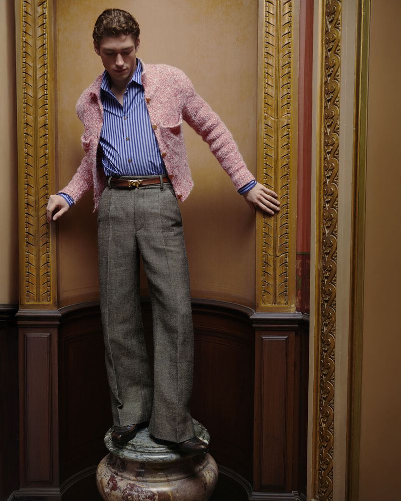 A young adult man in a pink jacket, striped shirt, and grey slacks poses in front of a gold-framed wall on a marble base indoors.