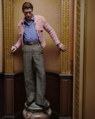 A young adult man in a pink jacket, striped shirt, and grey slacks poses in front of a gold-framed wall on a marble base indoors.