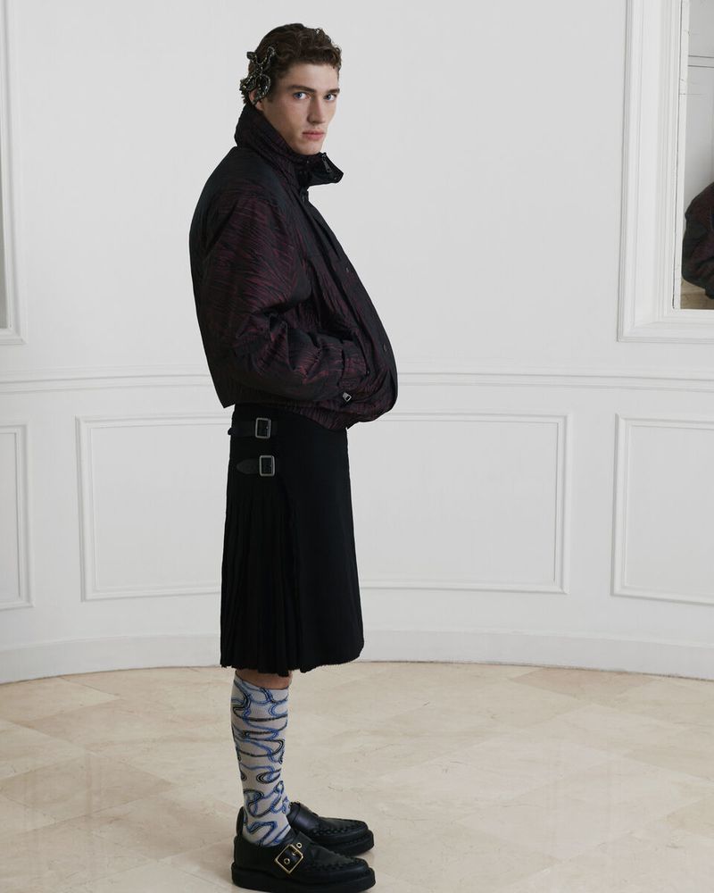 A fair-skinned male model poses indoors wearing a black and red jacket, black kilt, patterned socks, and black shoes, in a fashion studio setting.