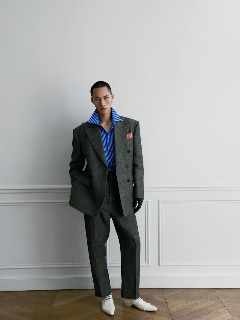 Male fashion model poses in a grey suit with a blue-striped collared shirt, white shoes, and black gloves against a white wall.