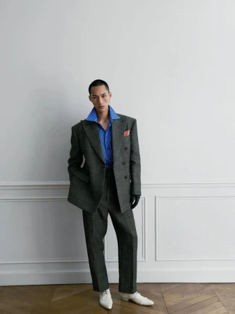 Model wearing a grey double-breasted suit and blue striped shirt against a white wall with wainscoting.