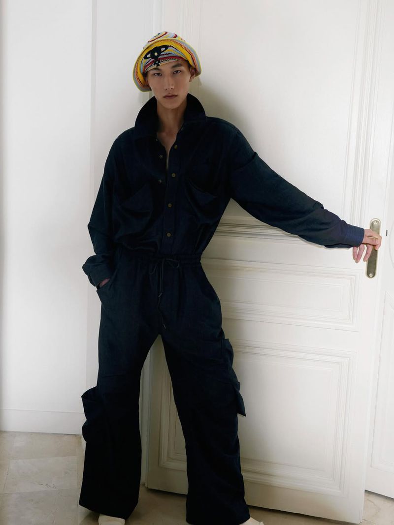 An adult, androgynous model wearing a jumpsuit and patterned headscarf, leaning against a white door. Fashion editorial photography.