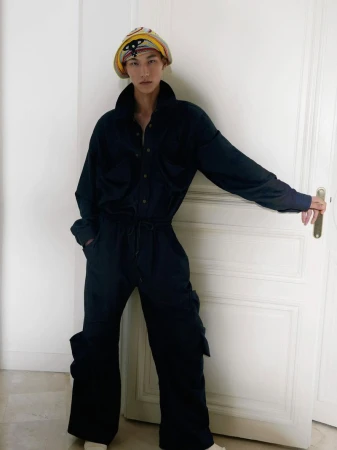 An adult, androgynous model wearing a jumpsuit and patterned headscarf, leaning against a white door. Fashion editorial photography.