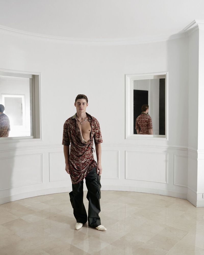 Young adult male in floral top and baggy pants poses in white interior space with mirrors, showcasing fashion and interior photography.