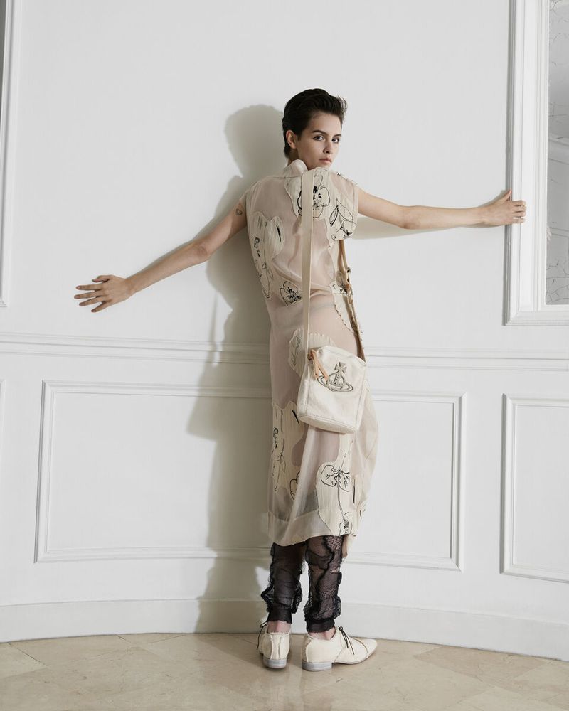 A slender woman in a printed dress and carrying a handbag looks back while standing indoors against a white wall with molding.