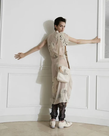 A slender woman in a printed dress and carrying a handbag looks back while standing indoors against a white wall with molding.