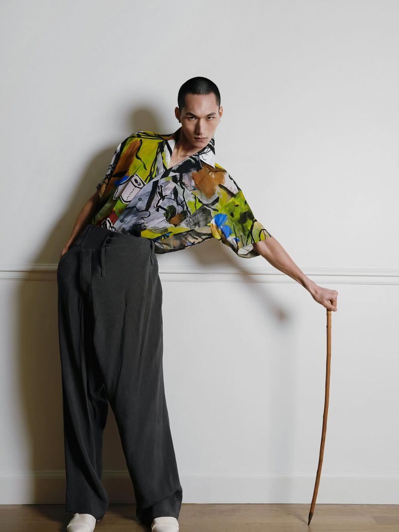 Adult male poses in an abstract-print short-sleeve shirt with dark wide-leg pants, white shoes, and a cane against a white wall.