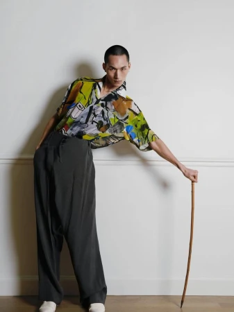 Adult male poses in an abstract-print short-sleeve shirt with dark wide-leg pants, white shoes, and a cane against a white wall.